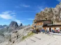 Rifugio alpino con gente seduta all'esterno, circondato da rocce imponenti e montagne. Un luogo ideale per le 12 escursioni da fare in Val di Fassa.