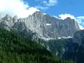 Imponente parete rocciosa del monte Civetta, con foreste di conifere ai suoi piedi e nuvole bianche nel cielo azzurro.