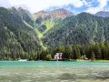 Un incantevole **Antholzer See** con acque turchesi, circondato da fitti boschi e imponenti montagne. Una casa bianca e un piccolo rifugio si affacciano sul lago.