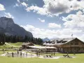 Un rifugio alpino con tavoli all'aperto e un laghetto dove giocano bambini, incorniciato da boschi e montagne imponenti sui **piani di Pezzè**.