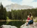 Mamma e figlia sedute su una roccia vicino a un lago alpino, con uno zaino blu. Un momento di relax per il loro lech dla lunch, con montagne imponenti sullo sfondo.