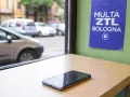 Un cellulare su un tavolo, con un cartello "Multa ZTL Bologna" appeso al muro. Fuori, auto in transito.