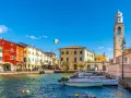 Un pittoresco porto dove si trova Lazise, con case colorate, barche e un campanile svettante sotto un cielo azzurro.