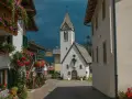 Un pittoresco vicolo a Rocca Pietore, con case fiorite e una chiesa dal cielo tempestoso.