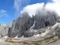 Rifugio in pietra ai piedi di imponenti vette rocciose, con neve residua e cielo azzurro. Un luogo incantevole al passo Valles.