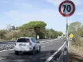 Un'auto bianca percorre una strada extraurbana con limite di 70 km/h. Alberi e cielo azzurro fanno da cornice.