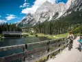 Famiglia in bicicletta sul ponte del **lago di San Vito**, con montagne imponenti e boschi sullo sfondo.