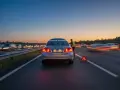 Auto in panne sul ciglio della strada, con un uomo in giubbotto catarifrangente e una segnalazione luminosa di pericolo. Traffico intenso al tramonto.