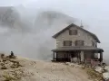 Rifugio Franz Kostner avvolto dalla nebbia, con un escursionista solitario su una panchina e le bandiere che sventolano.