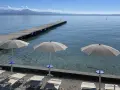 Ombrelloni e lettini al lido delle Bionde, con un pontile in legno che si estende sul lago limpido e montagne all'orizzonte.