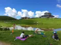 Persone rilassate sui prati verdi, vicino a un lago, sotto un cielo azzurro con nuvole bianche. Sembra un luogo ideale per i piani dell'avaro, godendosi la natura.