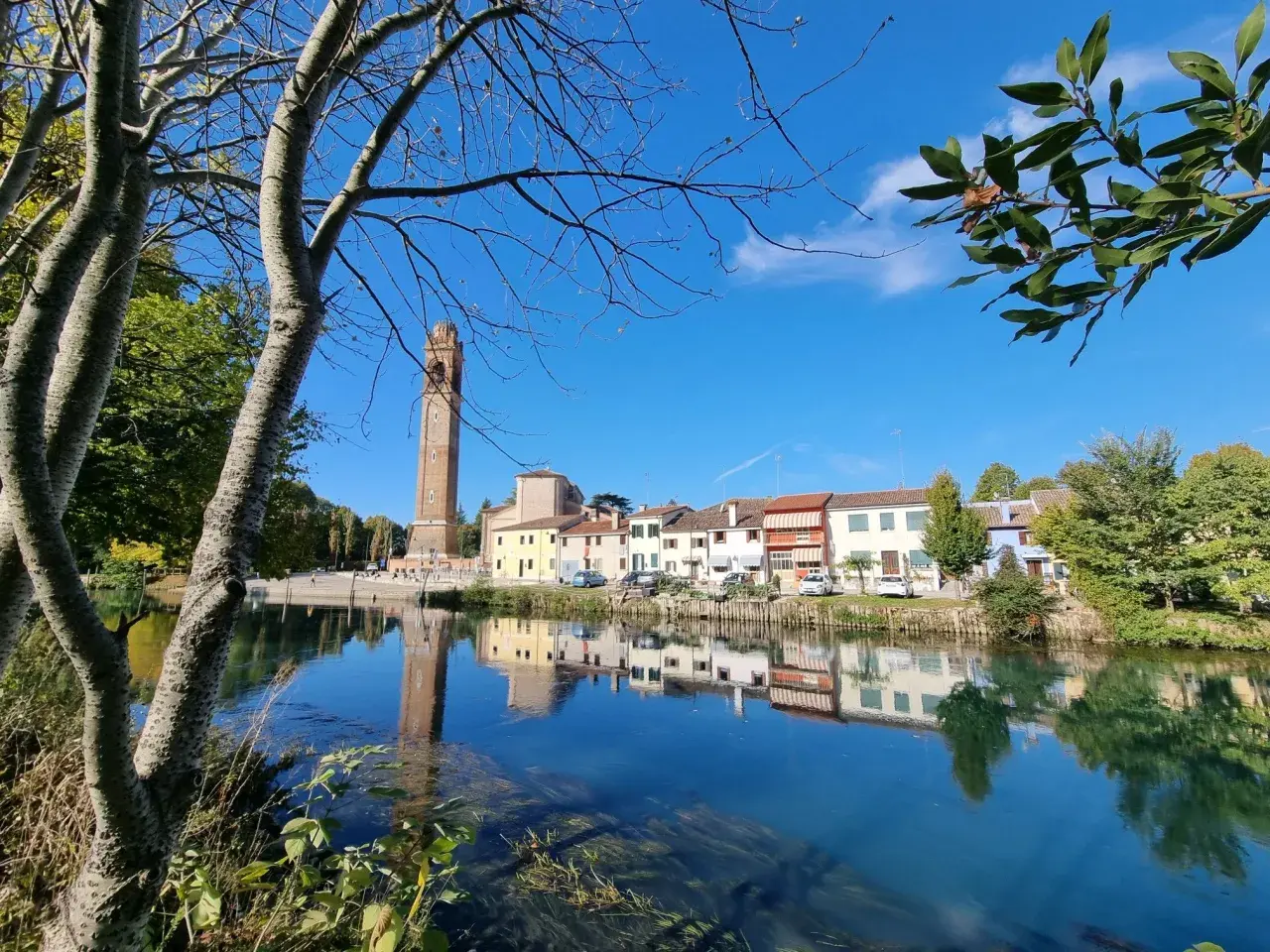 Un tranquillo fiume riflette le case colorate e l'alto campanile di Casale sul Sile, con alberi che incorniciano la scena.