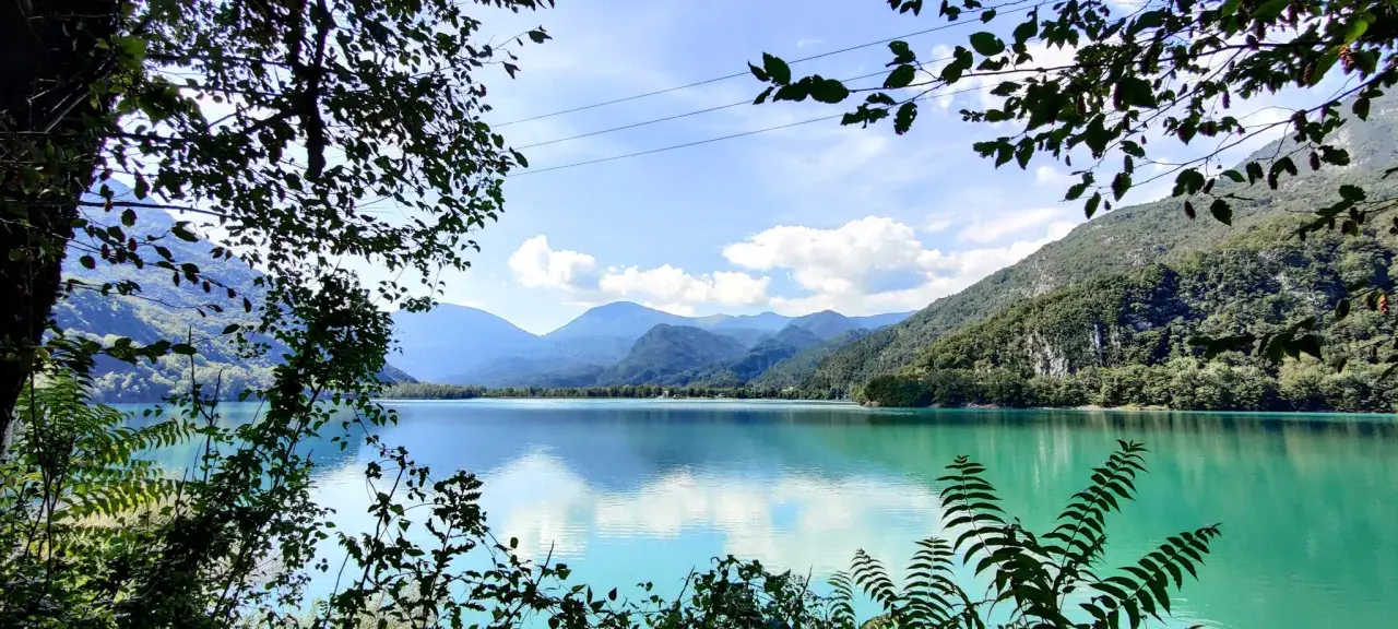 Un incantevole **lago di Cavazzo** con acque turchesi riflette le montagne verdi e il cielo azzurro, incorniciato da rigogliosa vegetazione.