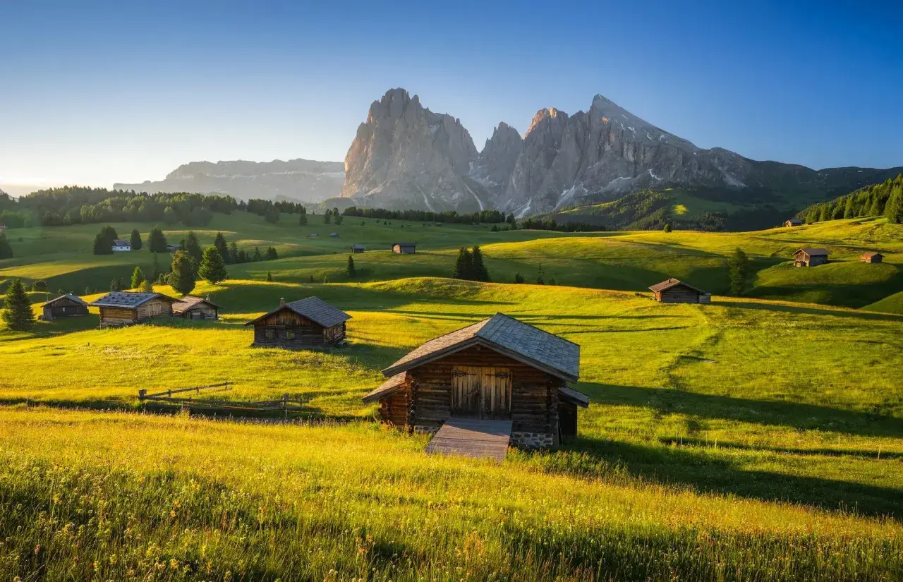 Alpe di Siusi al tramonto, con baite in legno su prati verdi e montagne imponenti sullo sfondo.