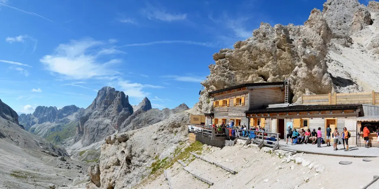Rifugio alpino con gente seduta all'esterno, circondato da rocce imponenti e montagne. Un luogo ideale per le 12 escursioni da fare in Val di Fassa.