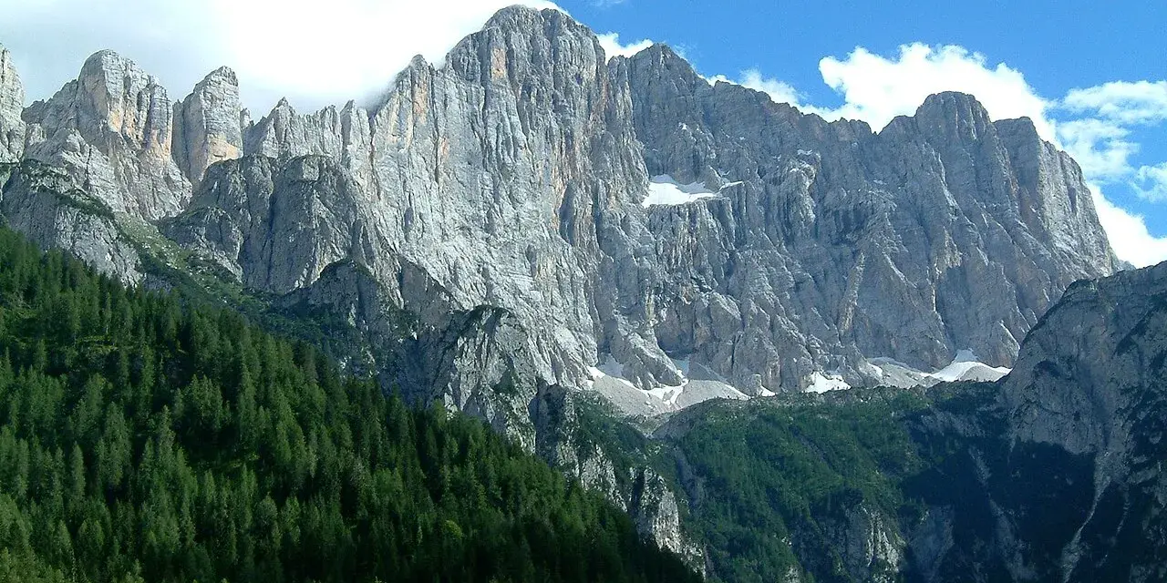 Imponente parete rocciosa del monte Civetta, con foreste di conifere ai suoi piedi e nuvole bianche nel cielo azzurro.