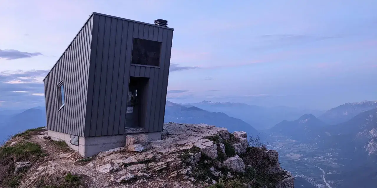 Il bivacco Giacomelli alla Madonnina, una struttura moderna su una cresta rocciosa, domina un paesaggio montano al tramonto.