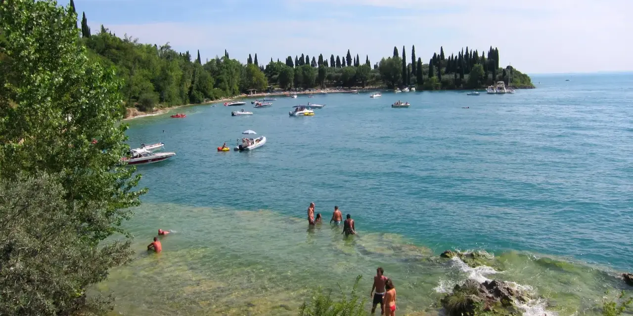 La baia delle Sirene a Garda, con gente che nuota e barche sull'acqua cristallina, sotto un cielo azzurro.