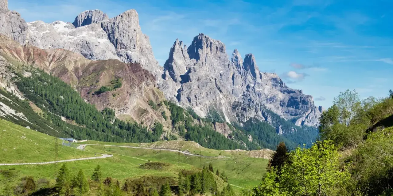 Strada panoramica verso le Pale di San Martino, con verdi pascoli e boschi che conducono alle maestose vette rocciose.