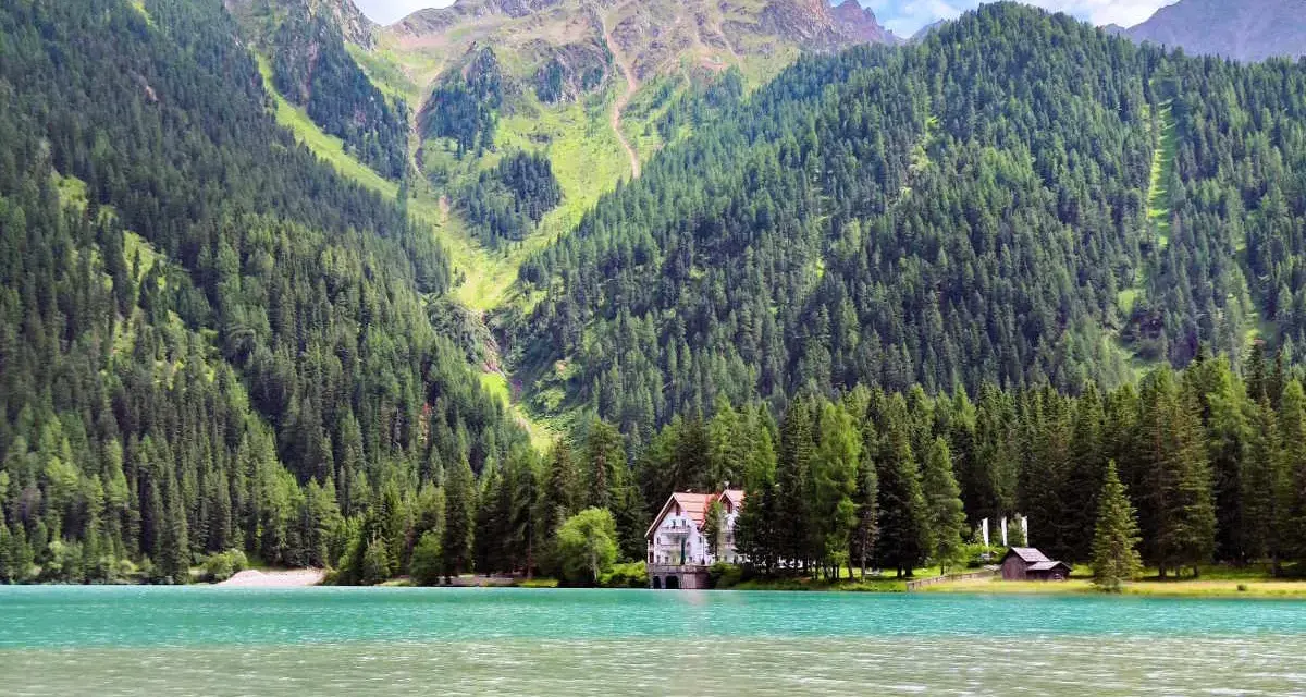 Un incantevole **Antholzer See** con acque turchesi, circondato da fitti boschi e imponenti montagne. Una casa bianca e un piccolo rifugio si affacciano sul lago.