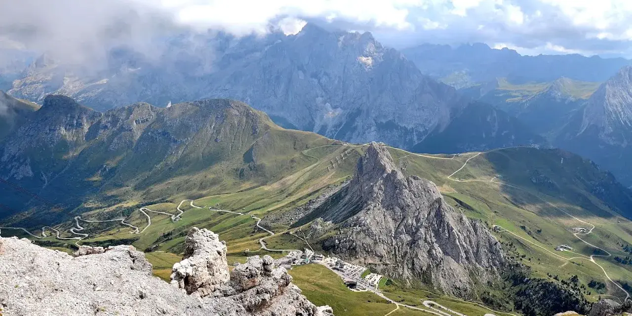 Panoramica mozzafiato del **Passo Pordoi** con strade tortuose che si snodano tra verdi pendii e imponenti vette rocciose, sotto un cielo nuvoloso.