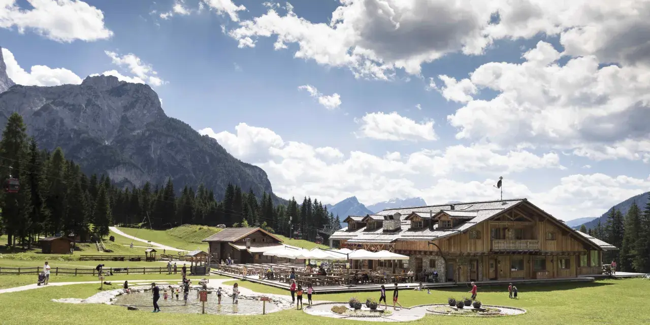 Un rifugio alpino con tavoli all'aperto e un laghetto dove giocano bambini, incorniciato da boschi e montagne imponenti sui **piani di Pezzè**.