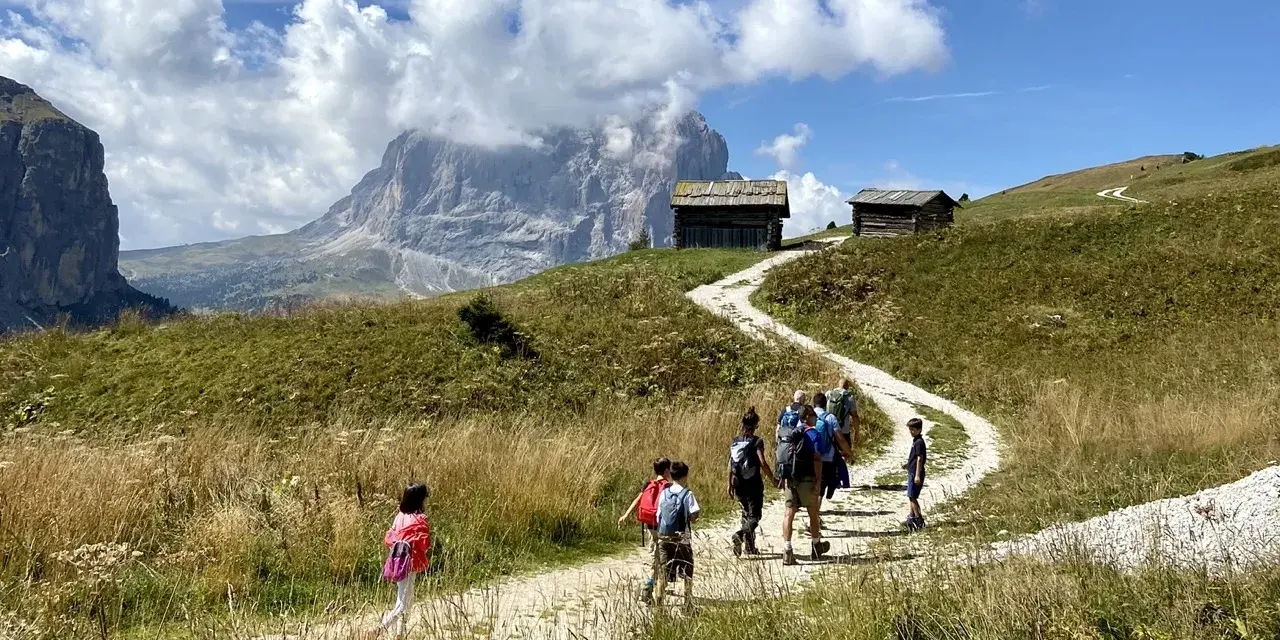 Famiglia in escursione su sentiero facile a Selva di Val Gardena, con baite e montagne sullo sfondo.