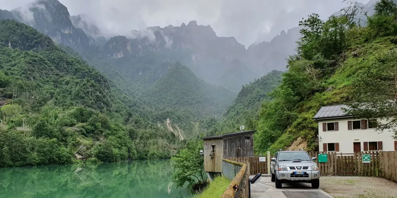 Il lago della Stua, incastonato tra montagne nebbiose e foreste lussureggianti, con una casa e un'auto parcheggiata vicino.