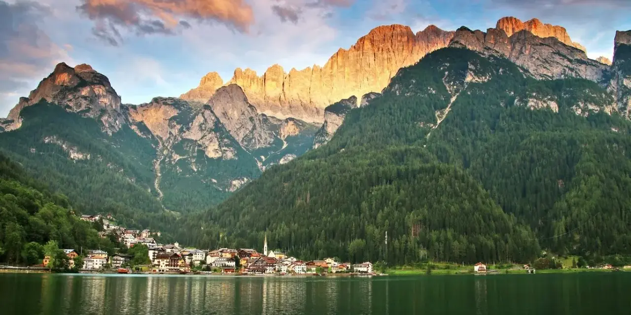 Un incantevole giro del lago di Alleghe, con il paese incastonato tra boschi e montagne maestose illuminate dal sole al tramonto.