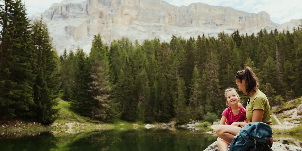 Mamma e figlia sedute su una roccia vicino a un lago alpino, con uno zaino blu. Un momento di relax per il loro lech dla lunch, con montagne imponenti sullo sfondo.