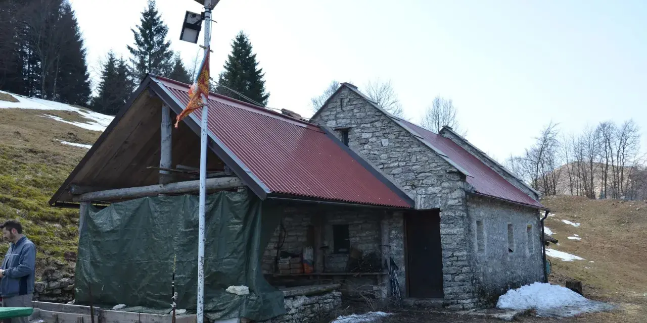 Bivacco Vallon Scuro: rifugio in pietra con tetto rosso, pannelli solari e telo verde. Un uomo si ripara dal freddo.