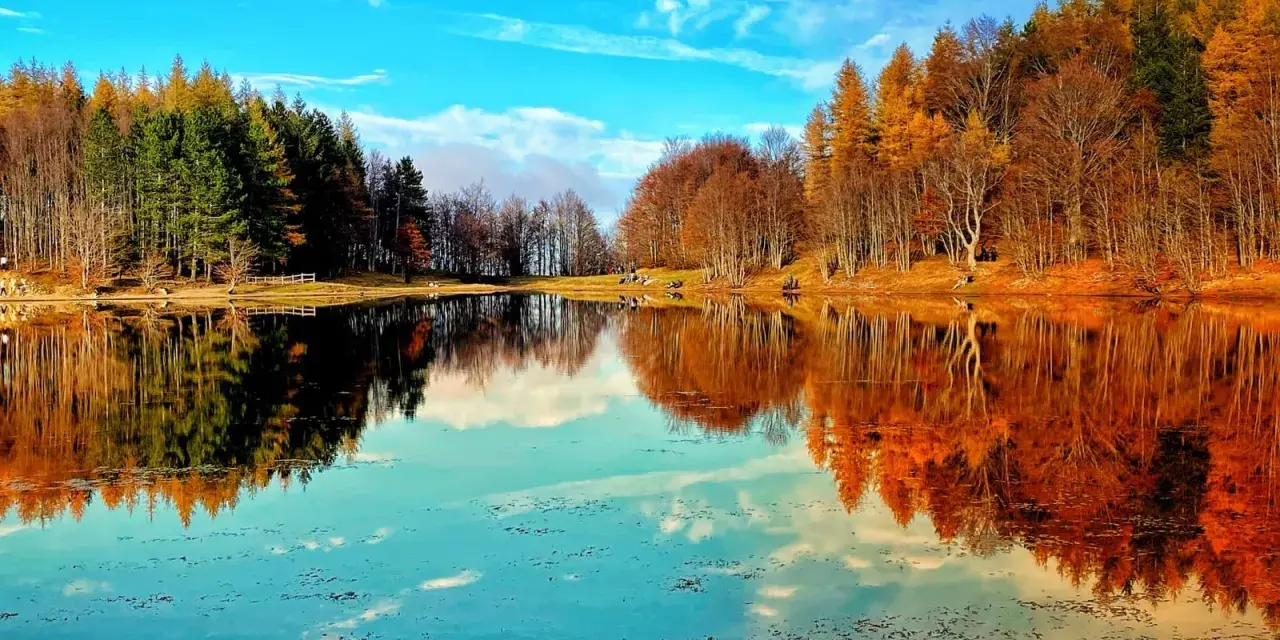 Specchio d'acqua sereno riflette alberi autunnali e cielo azzurro. Un angolo di pace tra i laghi dell'Emilia Romagna.