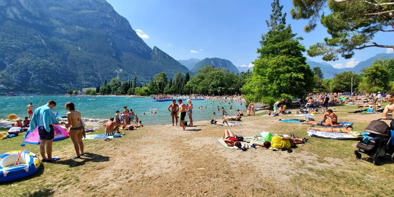 Spiaggia sabbioni Riva del Garda affollata di bagnanti sotto le montagne.