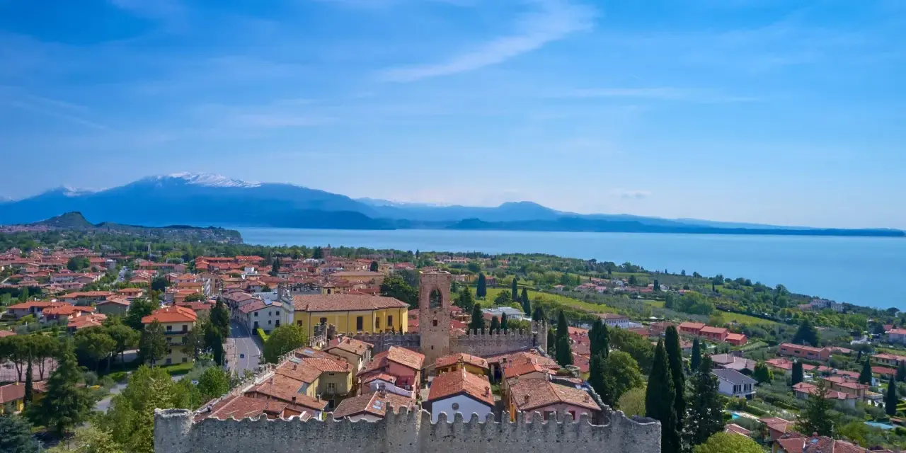 Vista aerea di Moniga del Garda, con il castello medievale in primo piano, il lago e le montagne sullo sfondo.