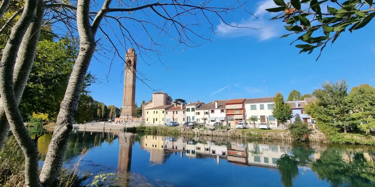 Un tranquillo fiume riflette le case colorate e l'alto campanile di Casale sul Sile, con alberi che incorniciano la scena.