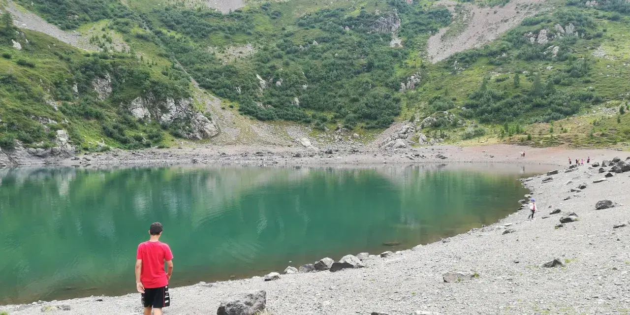 Ragazzo in maglietta rossa ammira il **lago di Erdemolo**, circondato da verdi pendii montuosi e rocce.