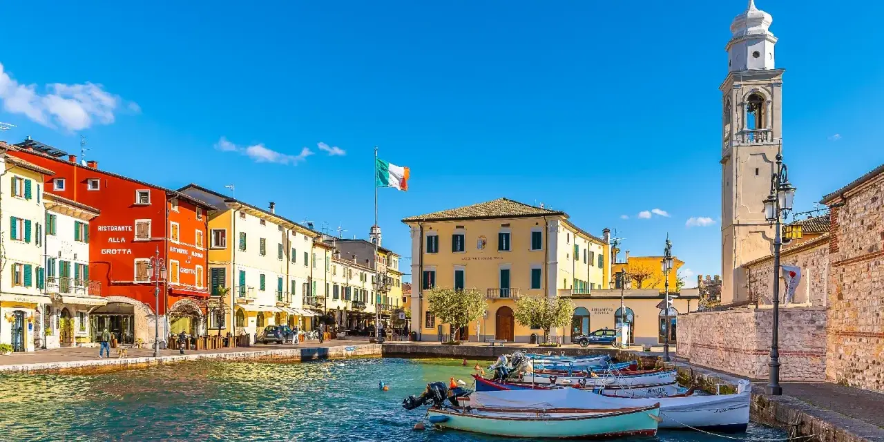 Un pittoresco porto dove si trova Lazise, con case colorate, barche e un campanile svettante sotto un cielo azzurro.