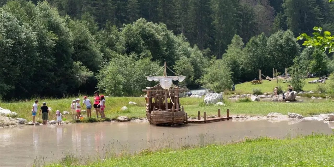 Bambini giocano vicino a un galeone di legno in un lago, mentre altri si rilassano sull'erba. Perfetto per le **san vigilio di marebbe passeggiate** estive.