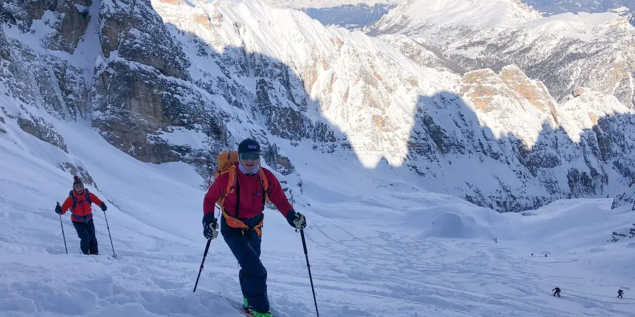 Sciatori scendono da un ripido pendio innevato sul Monte Cristallo, con vette maestose sullo sfondo. La **funivia** è lontana, ma l'avventura è appena iniziata.