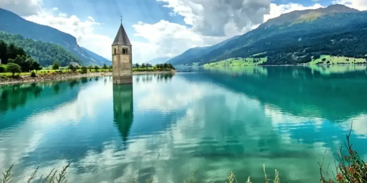 Torre sommersa nel lago di Resia, con montagne verdi e cielo nuvoloso.