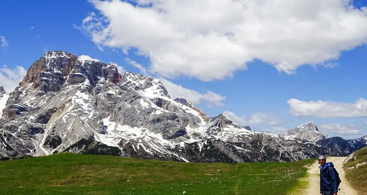Un uomo con bastoncini da trekking su un sentiero in Trentino, con imponenti montagne innevate sullo sfondo. Un'esperienza di trekking trentino facile e panoramica.