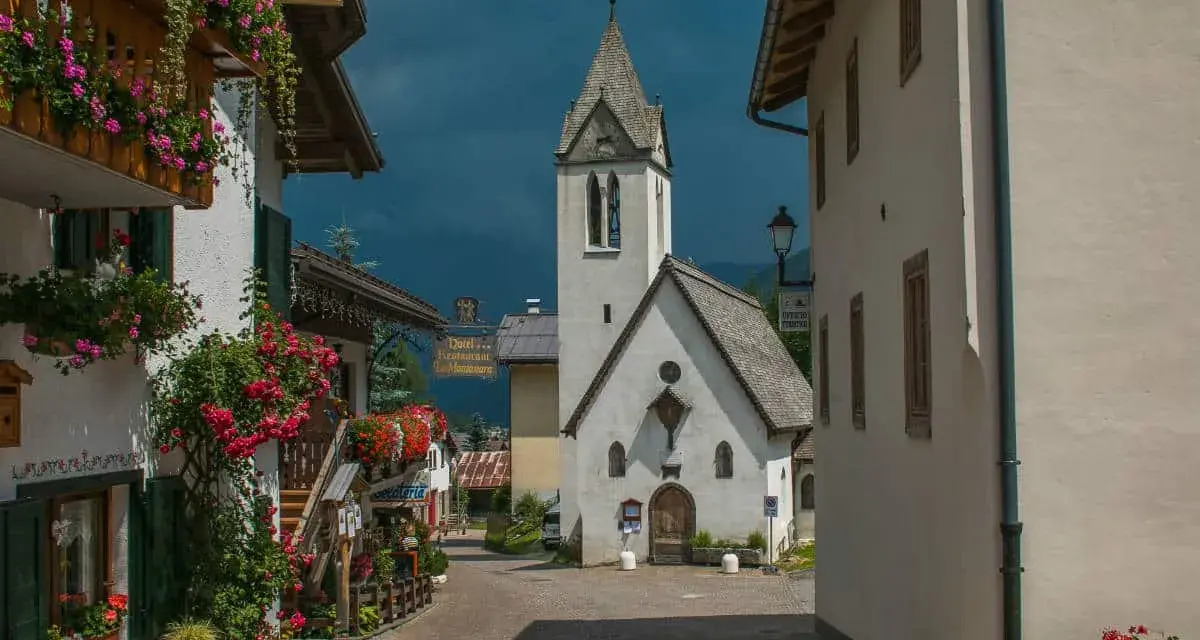 Un pittoresco vicolo a Rocca Pietore, con case fiorite e una chiesa dal cielo tempestoso.
