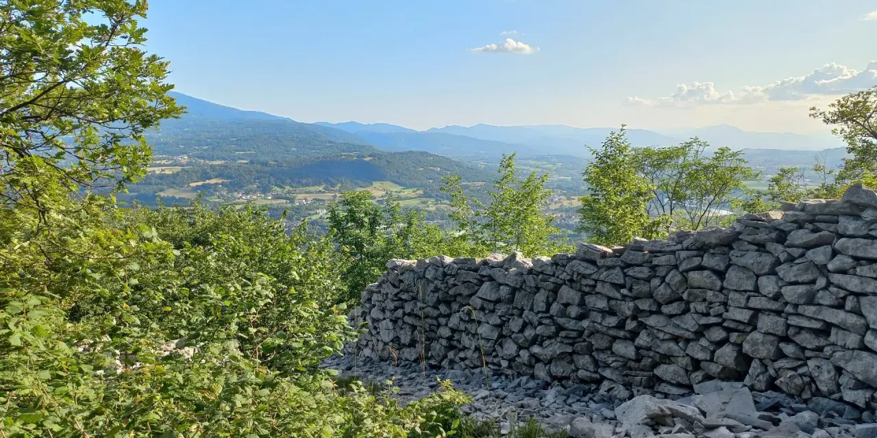 Muro di pietra a secco con vista panoramica su una valle alpina e montagne all'orizzonte.