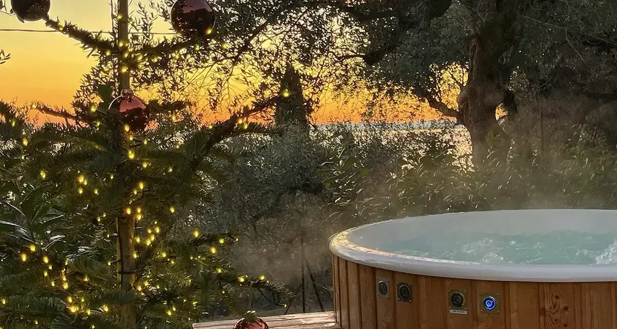 Un albero di Natale illuminato e una vasca idromassaggio fumante al tramonto sul Lago di Garda. Il camping è aperto tutto l'anno, un'oasi di relax invernale.