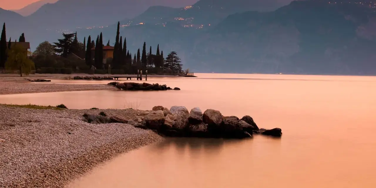 Brenzone sul Garda: cosa vedere? Spiaggia di ciottoli, cipressi e montagne al tramonto sul lago.