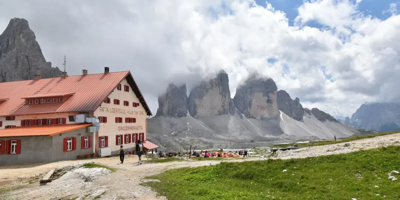 Il rifugio Locatelli, con le Tre Cime di Lavaredo avvolte dalle nuvole, accoglie escursionisti su un prato verde.