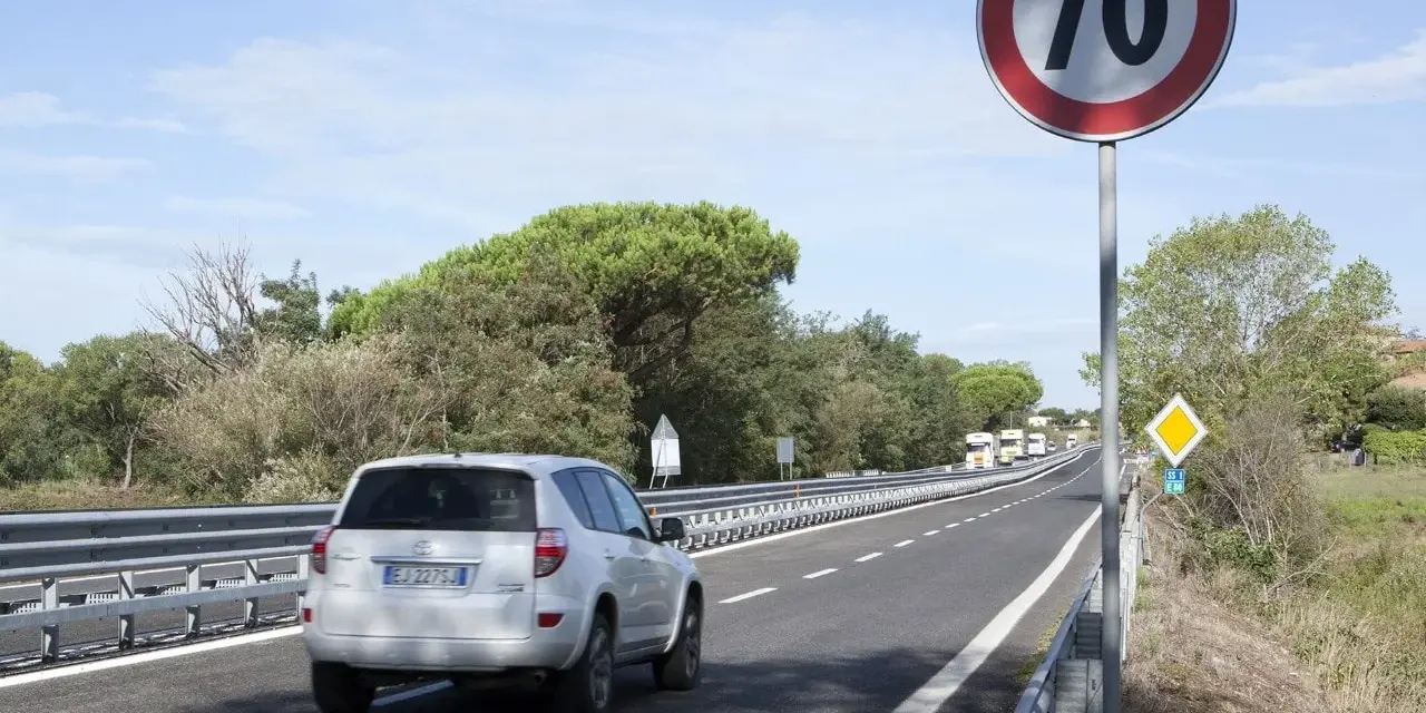 Un'auto bianca percorre una strada extraurbana con limite di 70 km/h. Alberi e cielo azzurro fanno da cornice.