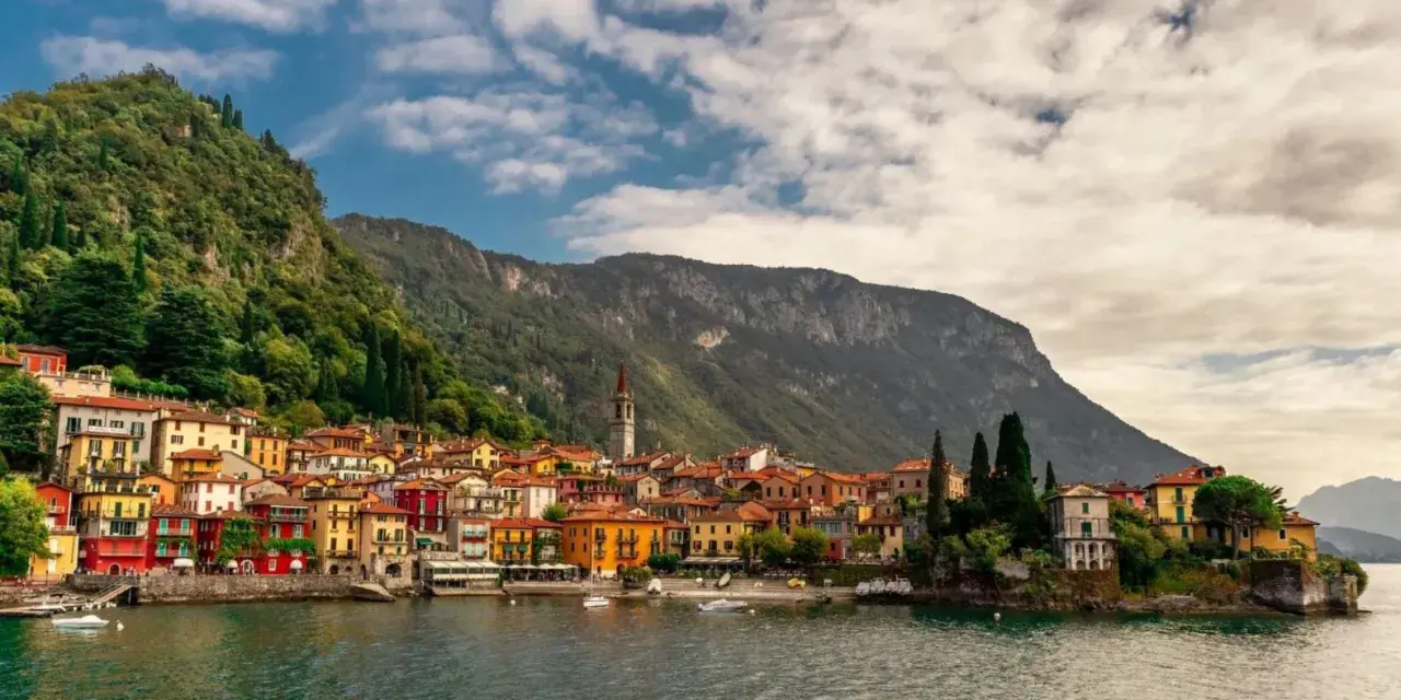 Colorate case affacciate sul lago di Lecco, con montagne verdi sullo sfondo e un cielo nuvoloso.