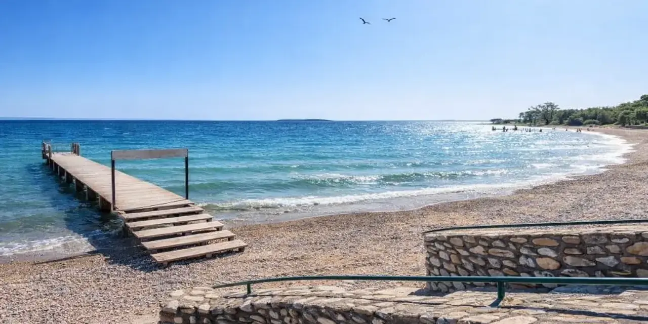 Molo in legno sul **Manerba del Garda strand**, con acqua cristallina e cielo azzurro. Due gabbiani volano sopra il lago.