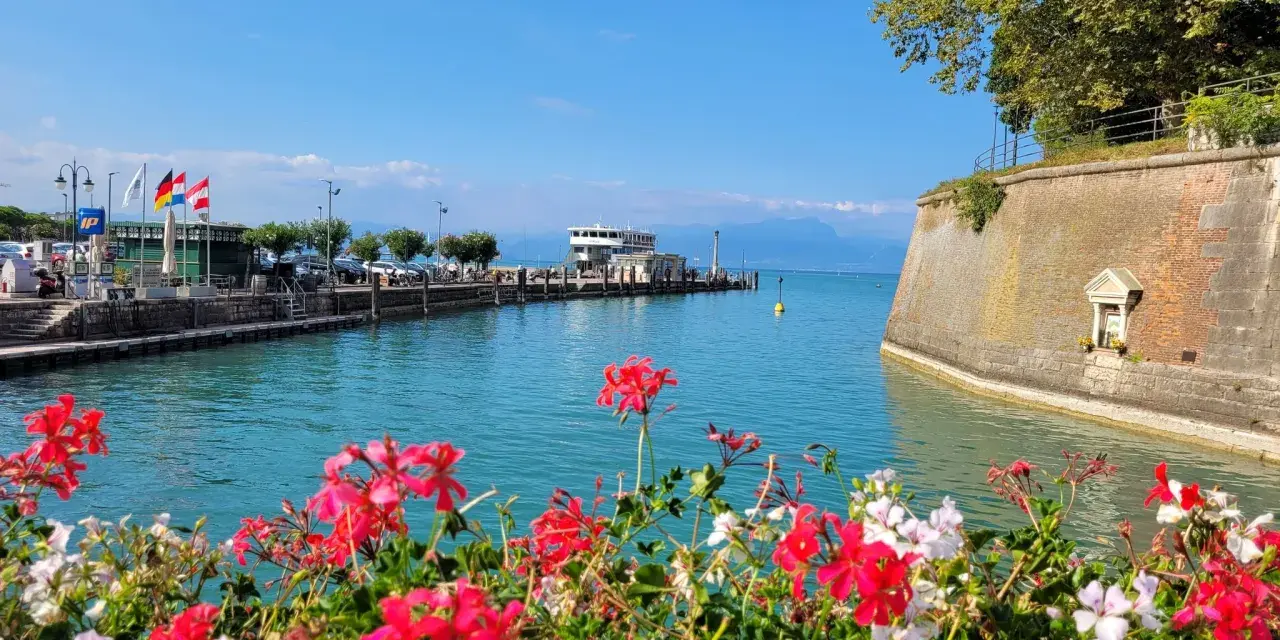 Fiori rossi e bianchi in primo piano, con un molo, un traghetto e montagne sullo sfondo. Un'immagine che evoca i paesi vicino Peschiera del Garda.
